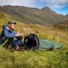 Man wearing a blue insulated jacket and cap sits on grass beside a small green bivvy tent, holding a mug and gazing across a mountainous valley under a partly cloudy sky.