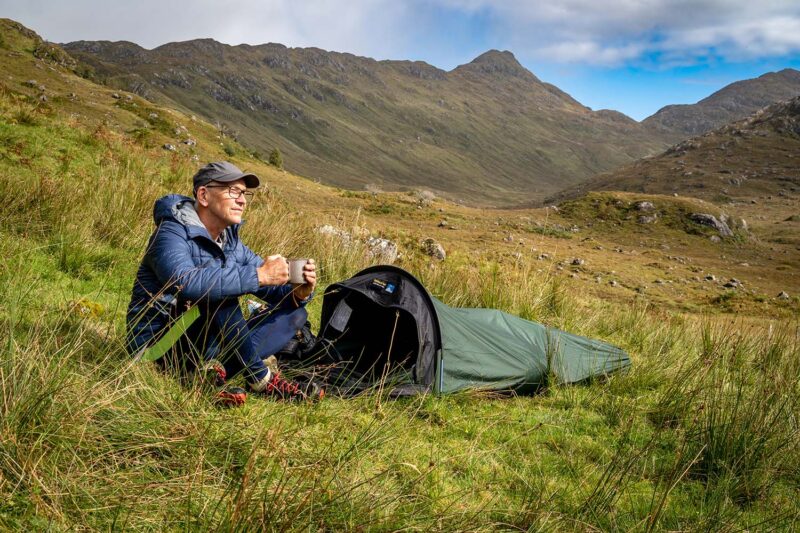 Man wearing a blue insulated jacket and cap sits on grass beside a small green bivvy tent, holding a mug and gazing across a mountainous valley under a partly cloudy sky.