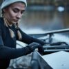 Young woman in a beanie and braided pigtails wearing a black thermal top and gloves adjusts the rigging on a rowing shell beside a calm river.