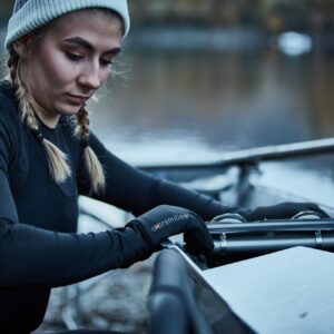 Young woman in a beanie and braided pigtails wearing a black thermal top and gloves adjusts the rigging on a rowing shell beside a calm river.