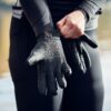 Close-up of a person pulling a textured black neoprene glove onto their hand while wearing a black wetsuit, with blurred water in the background.