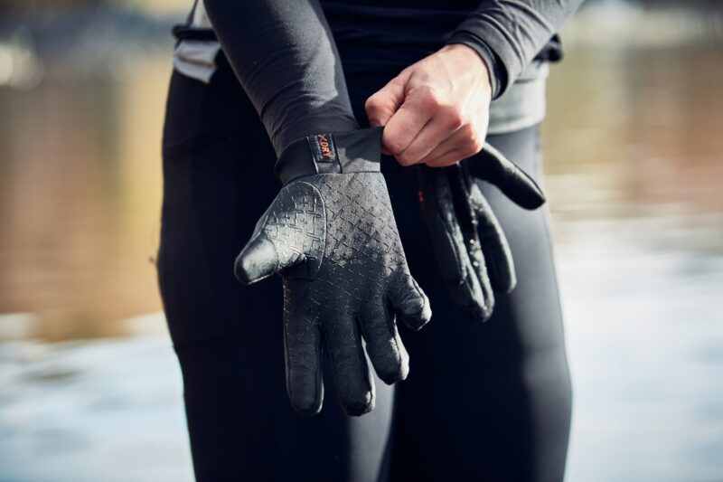 Close-up of a person pulling a textured black neoprene glove onto their hand while wearing a black wetsuit, with blurred water in the background.