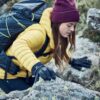 A hiker in a yellow down jacket and maroon beanie, wearing black gloves and a dark backpack, climbs over lichen-covered rocks and reaches forward with a gloved hand.