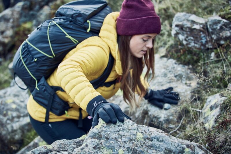 A hiker in a yellow down jacket and maroon beanie, wearing black gloves and a dark backpack, climbs over lichen-covered rocks and reaches forward with a gloved hand.
