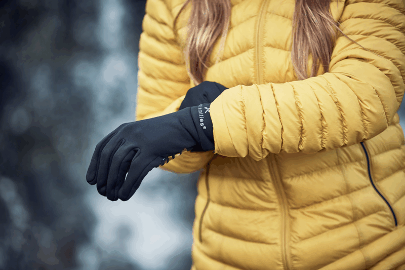 Close-up of a person in a yellow quilted jacket pulling on black thermal gloves against a blurred outdoor background.