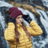 Person in a yellow puffer jacket and black gloves adjusting a maroon beanie with long hair and a focused expression, standing in front of a blurred waterfall.