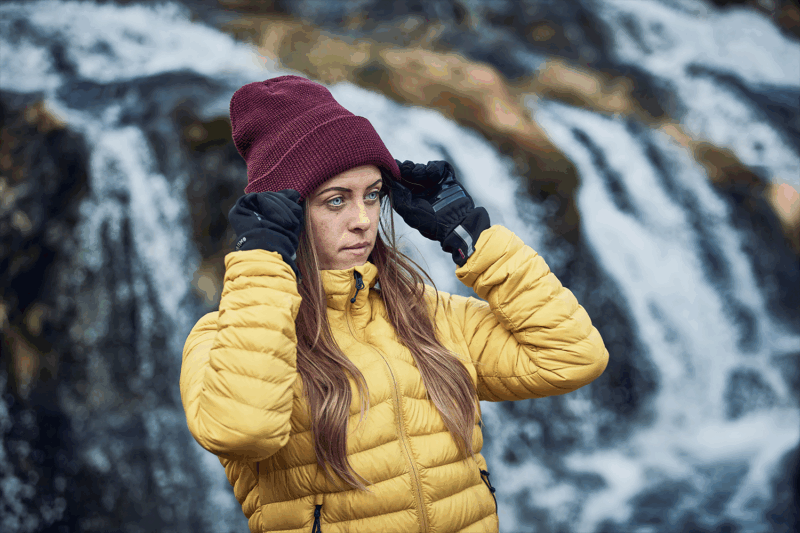 Person in a yellow puffer jacket and black gloves adjusting a maroon beanie with long hair and a focused expression, standing in front of a blurred waterfall.
