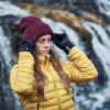 Woman in a yellow quilted jacket adjusting a maroon beanie with black gloves while standing in front of a blurred cascading waterfall
