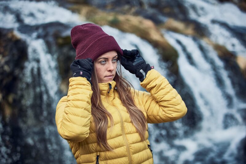 Woman in a yellow quilted jacket adjusting a maroon beanie with black gloves while standing in front of a blurred cascading waterfall
