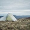 A pale green backpacking tent pitched on windswept grassy moorland overlooking rolling hills under a cloudy, overcast sky.