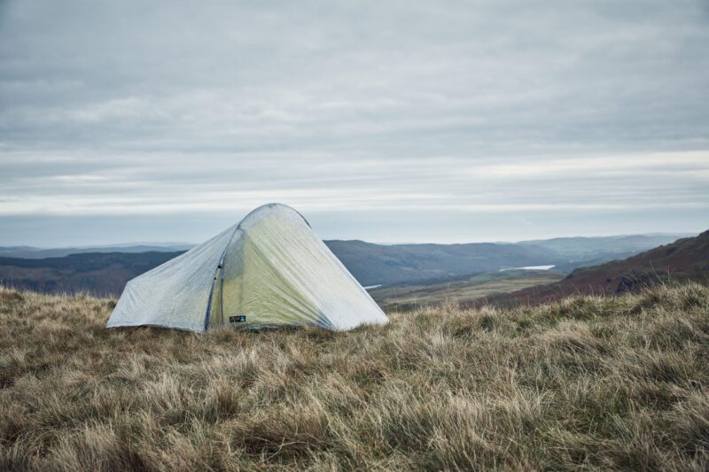 A pale green backpacking tent pitched on windswept grassy moorland overlooking rolling hills under a cloudy, overcast sky.