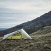 A lightweight tent with a yellow inner and translucent rainfly pitched on a windswept grassy hillside, steep rocky slopes rising behind it under a pale cloudy sky.