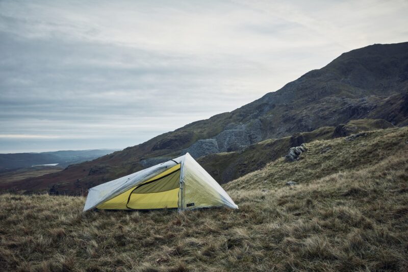 A lightweight tent with a yellow inner and translucent rainfly pitched on a windswept grassy hillside, steep rocky slopes rising behind it under a pale cloudy sky.