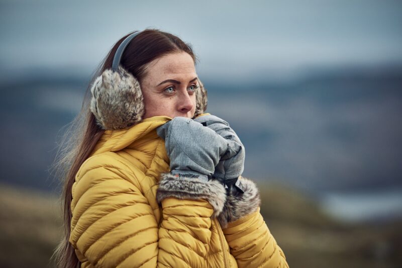 Woman in a yellow puffer jacket and fur earmuffs pressing grey, fur-trimmed mittens to her face while looking into a blurred cold outdoor landscape.