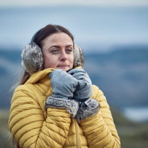 Woman in a yellow quilted jacket wearing grey furry earmuffs and grey mittens with fur trim, holding her hands near her face and gazing into the distance against a blurred cold, hilly coastal background.