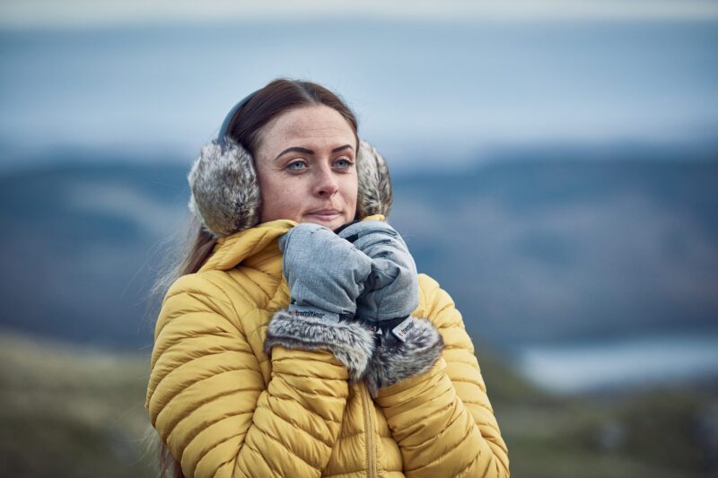 Woman in a yellow quilted jacket wearing grey furry earmuffs and grey mittens with fur trim, holding her hands near her face and gazing into the distance against a blurred cold, hilly coastal background.