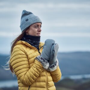Woman in a grey beanie and yellow puffer jacket with fur-lined grey mittens and a black neck gaiter, looking to the right against a soft-focus backdrop of hills and a cloudy sky.