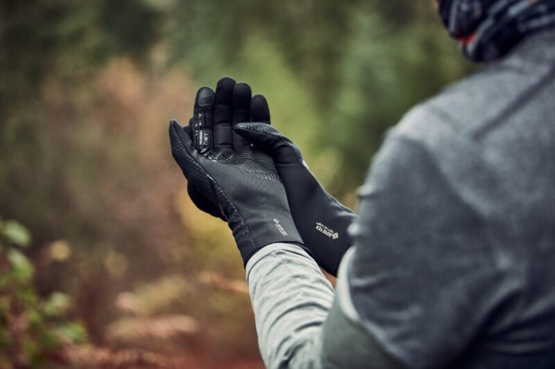 Hands of a person outdoors wearing black GORE‑TEX gloves held together as they adjust them, with a grey sleeve and a blurred green‑brown woodland background.