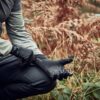 Cropped view of a person kneeling in an autumnal fern-filled wood, pulling on textured black grip gloves while wearing a grey long-sleeve top and black trousers.