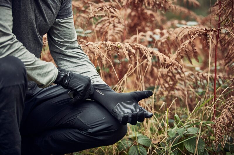 Cropped view of a person kneeling in an autumnal fern-filled wood, pulling on textured black grip gloves while wearing a grey long-sleeve top and black trousers.