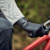 Close-up of a cyclist's gloved hand in a textured black glove gripping a mountain bike handlebar and brake lever, with a grey sleeve and red frame visible.