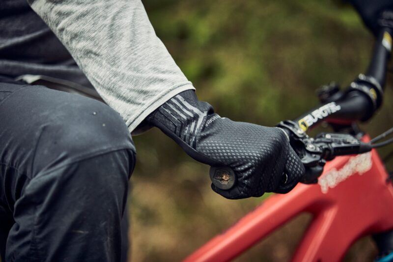 Close-up of a cyclist's gloved hand in a textured black glove gripping a mountain bike handlebar and brake lever, with a grey sleeve and red frame visible.