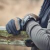 Close-up of a person's brown-and-black insulated gloves resting on a weathered, moss-covered wooden fence rail with a grey hoodie sleeve visible.