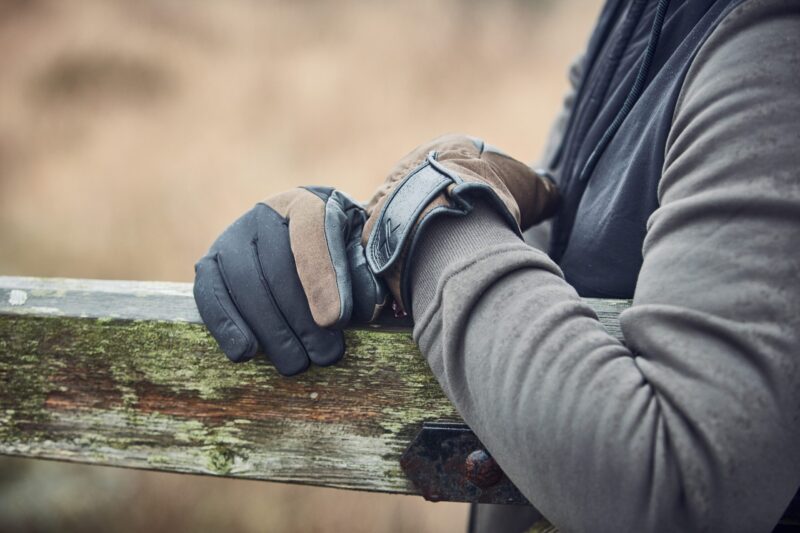 Close-up of a person's brown-and-black insulated gloves resting on a weathered, moss-covered wooden fence rail with a grey hoodie sleeve visible.