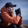 Man in an orange hooded jacket and navy cap sitting with a backpack, clasping his gloved hands near his face as he gazes thoughtfully across a distant landscape and blue sky.