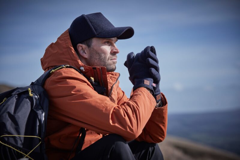 Man in an orange hooded jacket and navy cap sitting with a backpack, clasping his gloved hands near his face as he gazes thoughtfully across a distant landscape and blue sky.