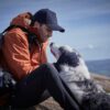 Man in an orange jacket and navy cap sits on a hillside with a backpack, leaning forward to cradle a fluffy white-and-grey dog with gloved hands as the dog lifts its head toward him against a distant blue-sky landscape.