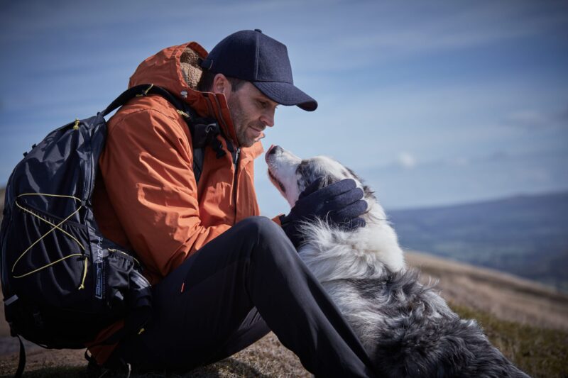Man in an orange jacket and navy cap sits on a hillside with a backpack, leaning forward to cradle a fluffy white-and-grey dog with gloved hands as the dog lifts its head toward him against a distant blue-sky landscape.
