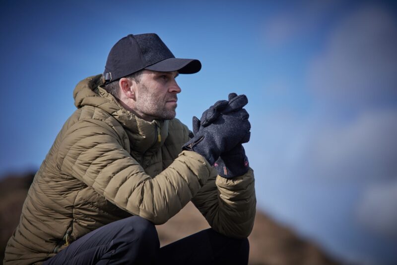 Man in an olive-green puffer jacket and black cap crouches on a rocky hillside, wearing dark gloves and clasping his hands as he gazes into the distance against a clear blue sky.