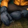 Close-up of a person sitting on grass in a mustard-yellow jacket wearing large black insulated mittens with adjustable straps and a visible GORE‑TEX tag, suggesting cold‑weather camping.