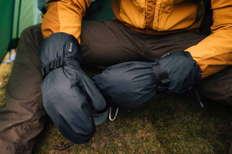 Close-up of a person sitting on grass in a mustard-yellow jacket wearing large black insulated mittens with adjustable straps and a visible GORE‑TEX tag, suggesting cold‑weather camping.