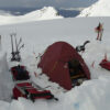A red camping tent set up in a snowy landscape, surrounded by ski equipment and sleds, with mountains and ocean visible in the background. Ideal for winter mountaineering and outdoor adventures.