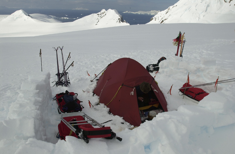 A red camping tent set up in a snowy landscape, surrounded by ski equipment and sleds, with mountains and ocean visible in the background. Ideal for winter mountaineering and outdoor adventures.