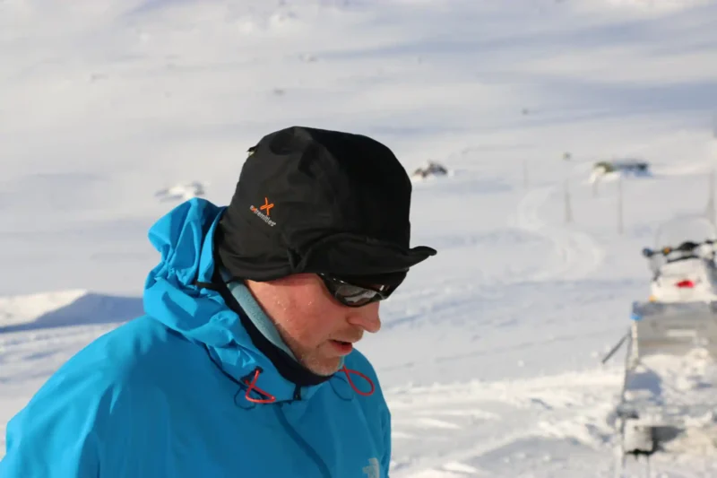 Man wearing a black winter hat and blue jacket in a snowy landscape, focused on a task, showcasing winter outdoor gear suitable for cold weather activities.