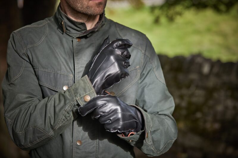 Close-up of a man’s torso and hands as he pulls on black leather gloves over the cuff of an olive-green waxed jacket, his stubbled chin visible and a blurred stone wall and grassy field in the background.