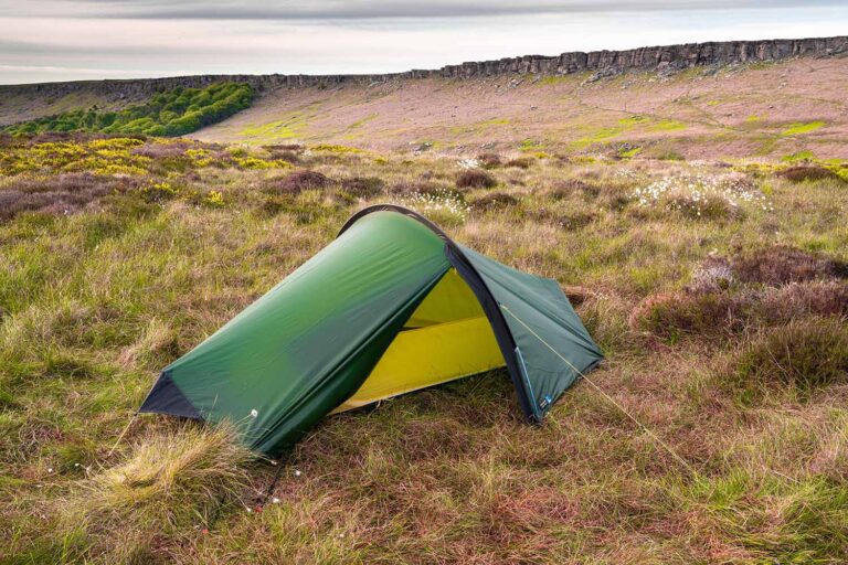 A small green two-person tent with a yellow inner lining staked on windswept heather and grass moorland dotted with white cotton-grass and yellow gorse, set against a low rocky escarpment and an overcast sky.