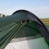 Close-up of a green camping tent's partially open entrance showing a dark rainfly with yellow toggles and a light grey inner panel, set against a grassy field and pale blue sky.