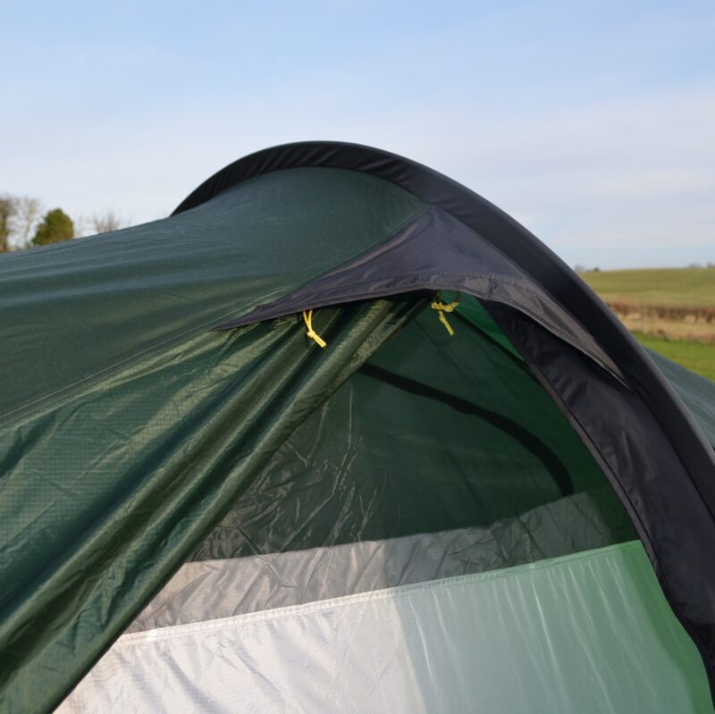 Close-up of a green camping tent's partially open entrance showing a dark rainfly with yellow toggles and a light grey inner panel, set against a grassy field and pale blue sky.