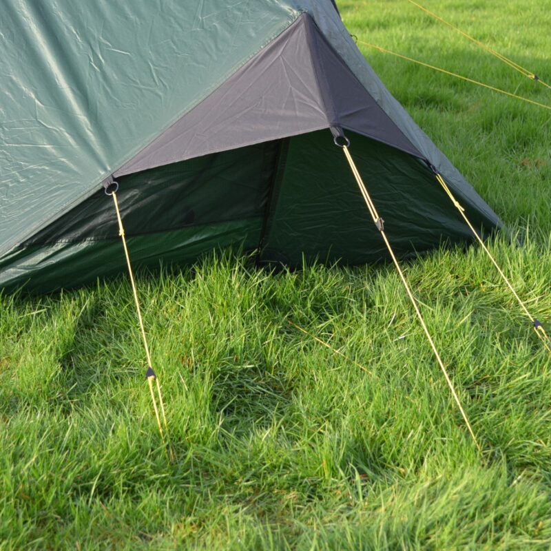 Close-up of a green tent's lower corner with a dark grey triangular vent flap held open by yellow guy lines and metal rings, staked into lush green grass.