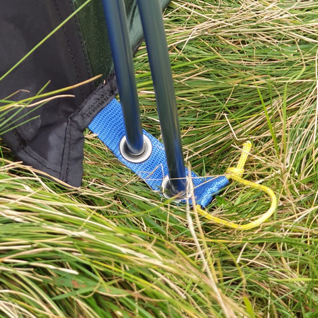 Close-up of two dark metal tent poles passing through a blue webbing loop with metal eyelets and a tied yellow cord, set among long green grass beside black fabric