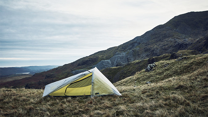 Lightweight yellow one-person tent pitched on a windswept grassy hillside with rocky mountains and an overcast sky in the background