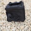 Small black padded pouch with a zippered top and Velcro strap, lying on pale loose gravel with a short looped side handle visible.