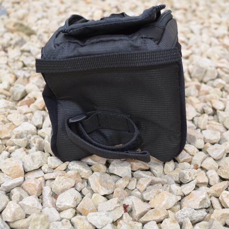 Small black padded pouch with a zippered top and Velcro strap, lying on pale loose gravel with a short looped side handle visible.