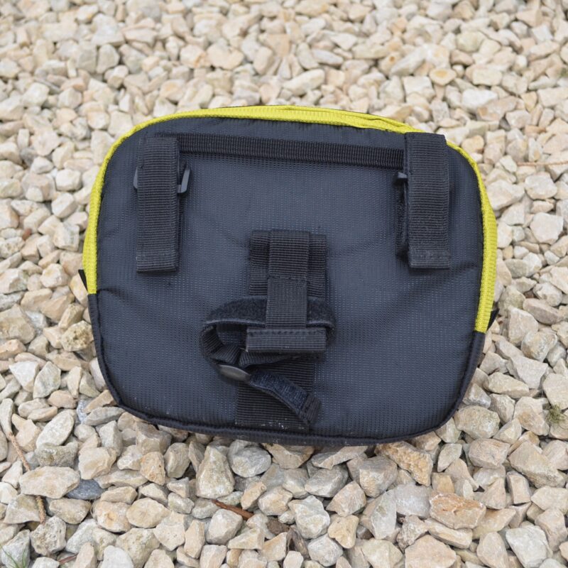 Small rectangular black fabric pouch with yellow trim, two vertical belt loops and a central velcro strap visible on its back, lying on a bed of pale gravel.