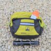 Yellow-green and grey Velo handlebar bag with an orange package peeking from its zippered pocket, resting on pale gravel with a small black bicycle pump placed in front.