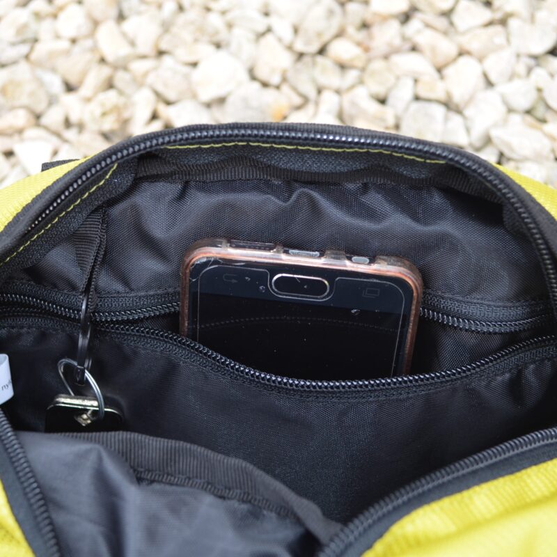 A black smartphone in a clear protective case sticks out of a zippered inner pocket of a yellow fabric pouch, a key on a ring peeks from the pocket and pale gravel is visible in the background.